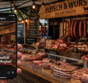 Eine Person hält ein Smartphone vor einen Metzgerei-Marktstand mit frischen Fleisch- und Wurstwaren. Auf dem Smartphone ist eine Instagram-Story des Standes mit dem Text „Beste Qualität aus der Region!“ zu sehen. Im Hintergrund herrscht eine lebendige Marktatmosphäre mit rustikalen Holzelementen und zahlreichen Fleischspezialitäten.